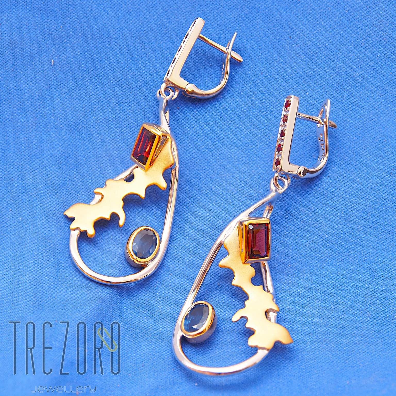 Adventure Road Designer Earrings. Sterling Silver with Garne,t Sapphire. Rhodium and Gold Plated. On blue.